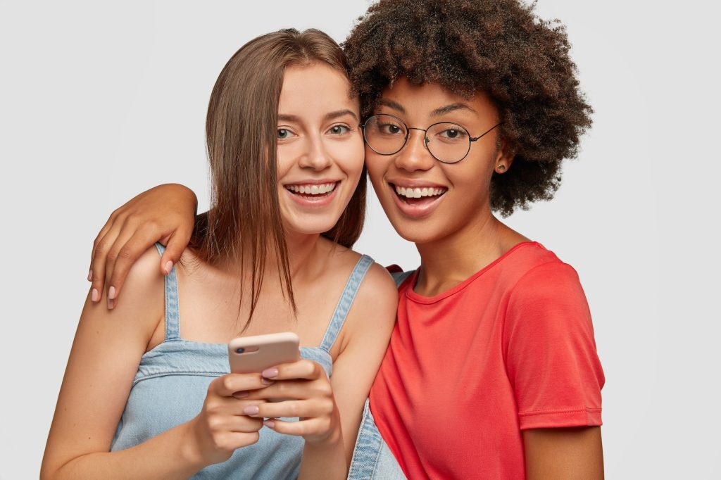 Diverse women with cheerful expressions read posts in social networks via cellular, hug and smile po