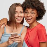 Diverse women with cheerful expressions read posts in social networks via cellular, hug and smile po