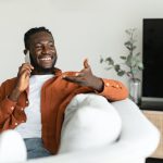 Making call. Happy african american man talking on smartphone and gesturing, resting on sofa at home