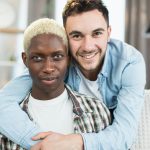 Two multicultural gays embracing and smiling on couch