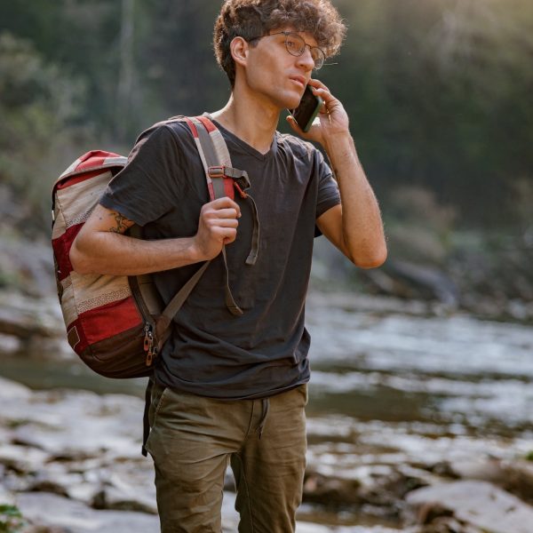 Lost young man tourist with backpack roaming at river in mountains and talking on cellphone.