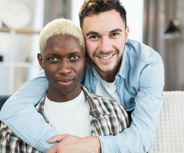 Two multicultural gays embracing and smiling on couch